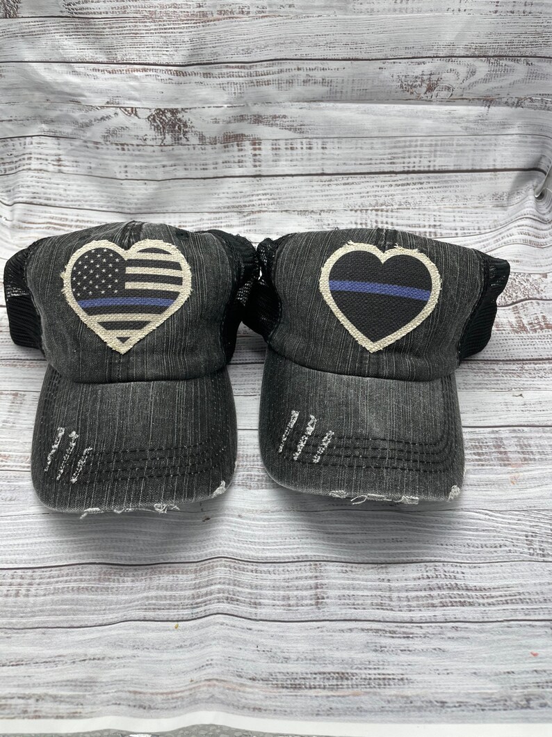 May include: Two black denim trucker hats with distressed detailing. One hat features a white heart with an American flag and a blue line, the other hat features a white heart with a blue line.