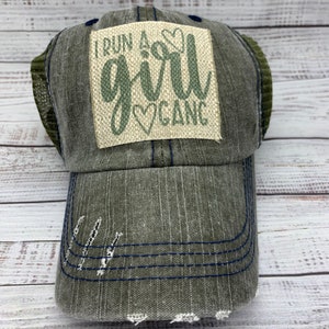 May include: A green and blue distressed trucker hat with a patch that reads "I run a girl gang" in green lettering with a heart design.