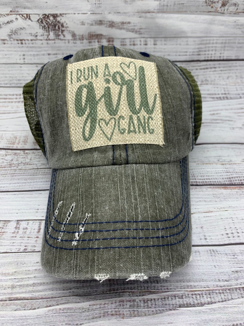 May include: A green and blue distressed trucker hat with a patch that reads "I run a girl gang" in green lettering with a heart design.