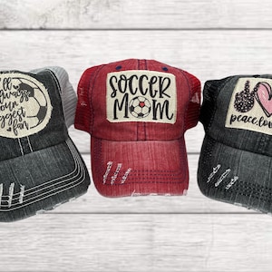 May include: Three black and red distressed trucker hats with embroidered soccer-themed patches. The patches say "I'll always be your biggest fan", "Soccer Mom", and "Peace Love Soccer".