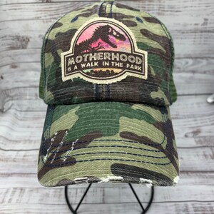 May include: A green camouflage trucker hat with a distressed look. The hat features a patch with a dinosaur silhouette and the text "MOTHERHOOD IS A WALK IN THE PARK".