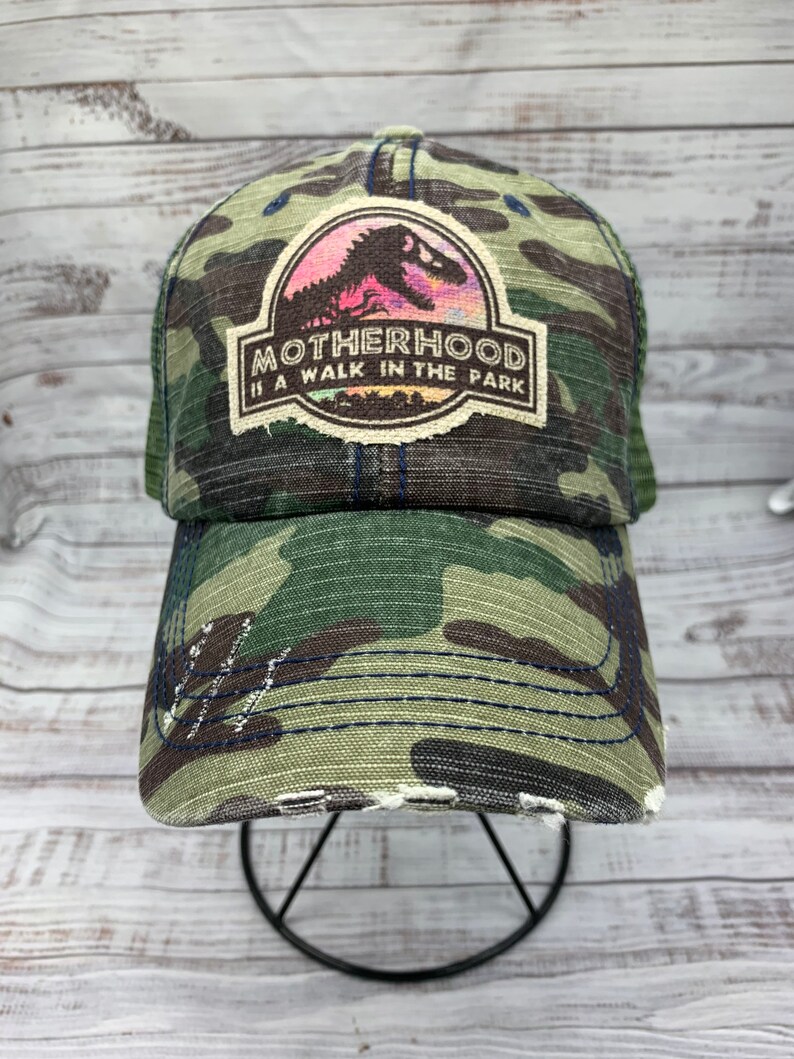 May include: A green camouflage trucker hat with a distressed look. The hat features a patch with a dinosaur silhouette and the text "MOTHERHOOD IS A WALK IN THE PARK".