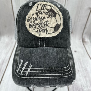 May include: A black and gray distressed trucker hat with a white mesh back. The front of the hat features a brown patch with a white soccer ball and the text "I'll always be your biggest fan".