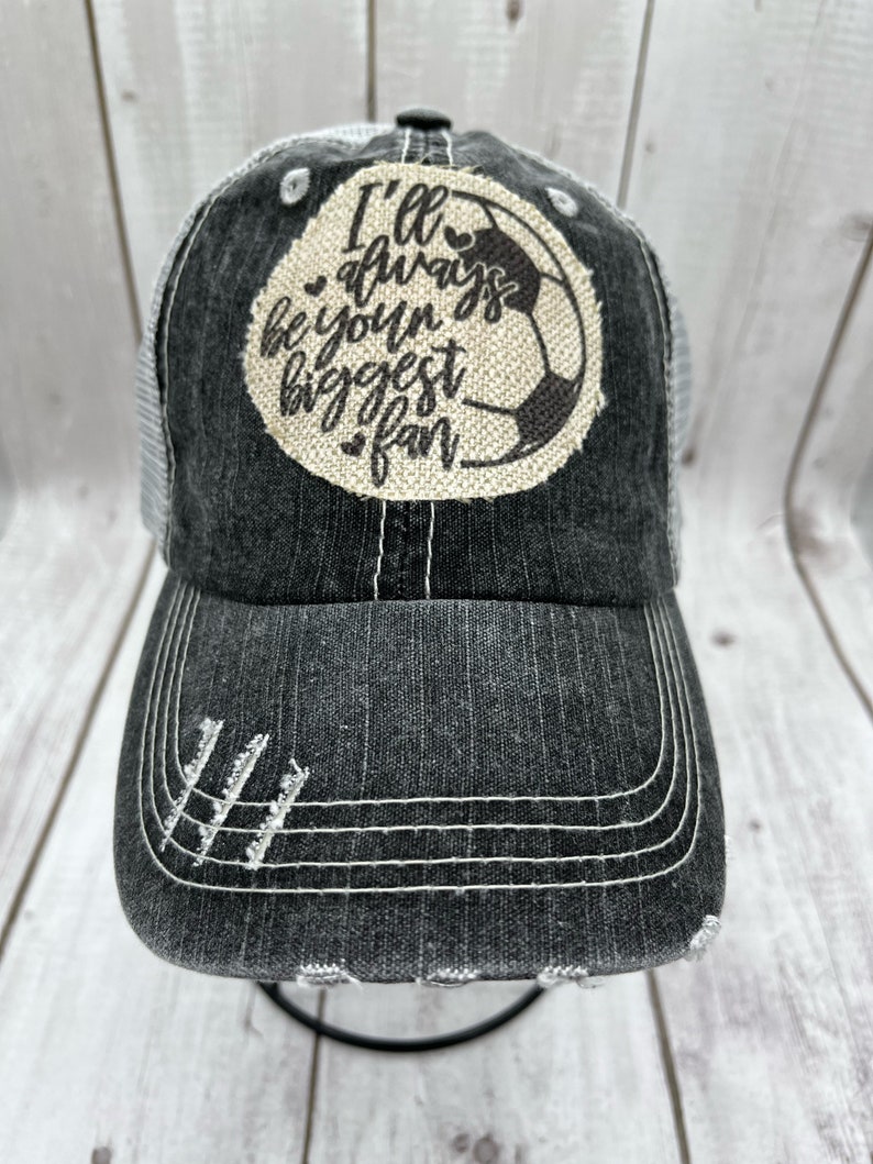 May include: A black and gray distressed trucker hat with a white mesh back. The front of the hat features a brown patch with a white soccer ball and the text "I'll always be your biggest fan".