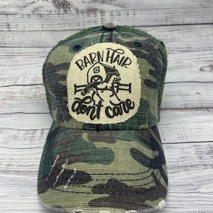 May include: A camouflage trucker hat with a distressed look. The hat has a patch on the front that says "Barn Hair don't care" with a drawing of a horse and barn.