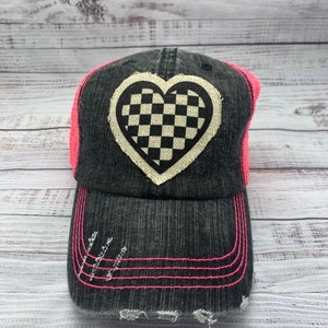 May include: A black denim trucker hat with a pink mesh back. The hat has a distressed look and features a white checkered heart patch.