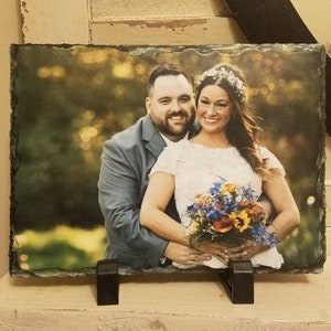 May include: A black slate photo holder with a printed image of a couple in a wedding setting. The couple is standing in a green grassy area with trees in the background. The woman is wearing a white dress and a flower crown. The man is wearing a gray suit. The couple is holding a bouquet of flowers.