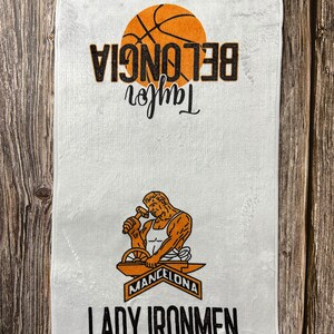 May include: White towel with an orange basketball and the text "Taylor Beloncia" in black. Below the basketball is a black and orange illustration of a person working on a metal anvil with the text "Mancelona Lady Ironmen" in black.