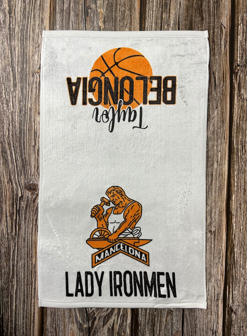 May include: White towel with an orange basketball and the text "Taylor Beloncia" in black. Below the basketball is a black and orange illustration of a person working on a metal anvil with the text "Mancelona Lady Ironmen" in black.