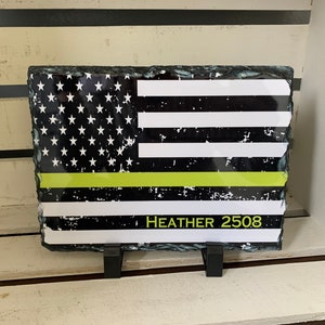 May include: A black and white American flag with a thin yellow line across the middle. The flag is printed on a slate stone with a rough, natural edge. The words "HEATHER 2508" are printed below the flag.