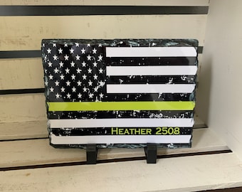 May include: A black and white American flag with a thin yellow line across the middle. The flag is printed on a slate stone with a rough, natural edge. The words "HEATHER 2508" are printed below the flag.