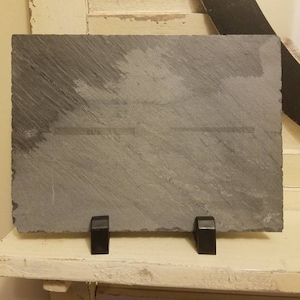 May include: A black slate plaque with a smooth, textured surface. The plaque is mounted on a black metal stand with two legs.