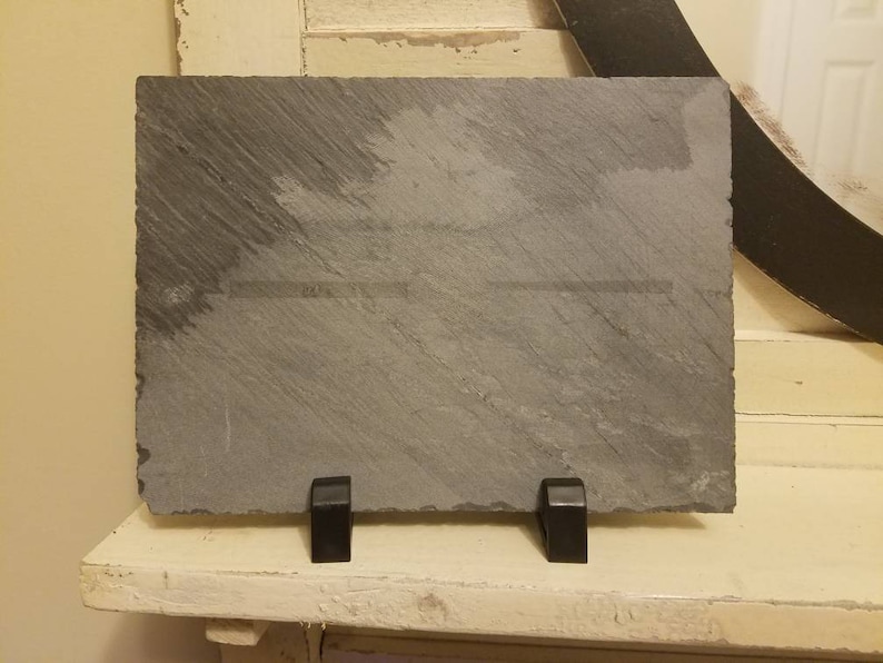 May include: A black slate plaque with a smooth, textured surface. The plaque is mounted on a black metal stand with two legs.