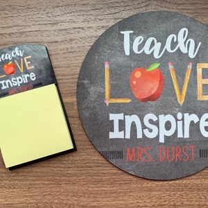 May include: A notepad and a round chalkboard-style sign with the words "Teach Love Inspire" and a red apple graphic. The notepad has yellow sticky notes. The sign also includes the name "MRS. DURST".
