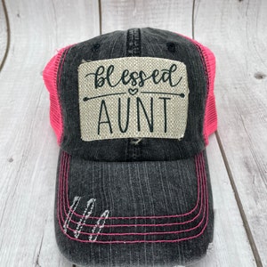 May include: A black and pink trucker hat with a distressed denim front and a pink mesh back. The hat has a patch on the front that says "blessed aunt" with a small heart symbol.