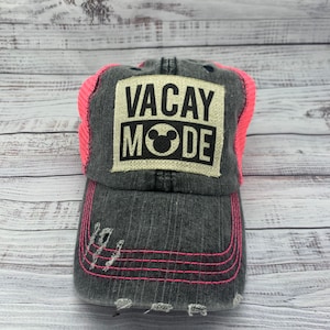 May include: A black and gray distressed baseball cap with a pink mesh back. The front of the cap has a white patch with the text "VACAY MODE" in black letters and a black Mickey Mouse head.