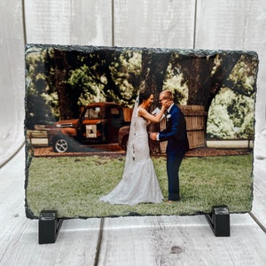 May include: A black slate photo frame with a rustic, uneven edge, displaying a color photograph of a bride and groom. The couple is standing in a grassy field with a vintage red truck in the background. The bride is wearing a white wedding dress and veil, and the groom is wearing a dark suit.