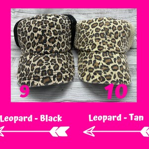 May include: Two leopard print trucker hats, one with a black mesh back and one with a tan mesh back. The hats are on a wooden surface. The text "Leopard - Black" and "Leopard - Tan" is below the hats.