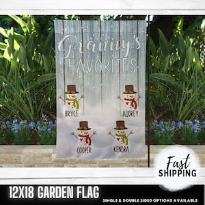 May include: A 12x18 inch garden flag with a winter theme. The flag features a white wooden background with a blue sky and falling snow. There are four snowmen with personalized names: Bryce, Audrey, Cooper, and Kendra. The text "Grammy's Favorites" is at the top of the flag.