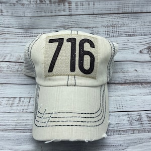 May include: A white mesh trucker hat with a distressed look and a black patch with the number 716 on it.