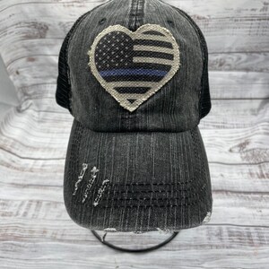 May include: Black distressed baseball cap with a white heart-shaped patch featuring an American flag with a blue line. The cap has a mesh back.