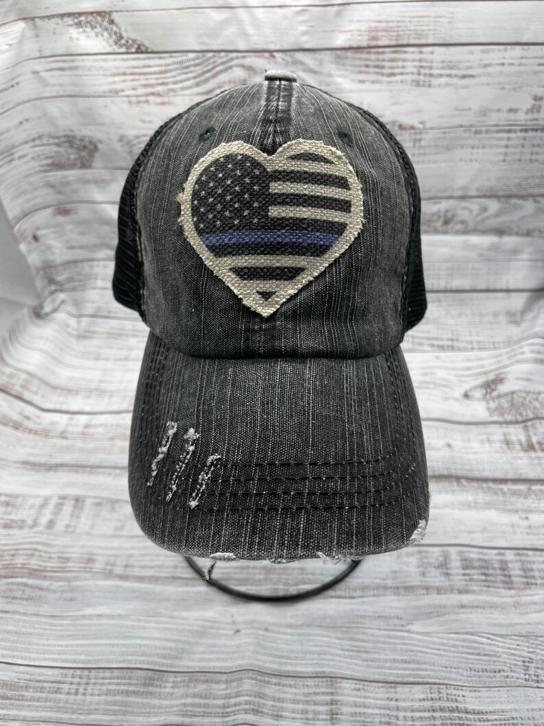 May include: Black distressed baseball cap with a white heart-shaped patch featuring an American flag with a blue line. The cap has a mesh back.