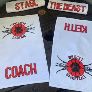 May include: Four white towels with red and black printed designs. One towel has the text "STAGL" printed in red. Another towel has the text "THE BEAST" printed in red. The third towel has the text "WILDCAT BASKETBALL" and a paw print inside a basketball. The fourth towel has the text "COACH" printed in red and the text "WILDCAT BASKETBALL" and a paw print inside a basketball.
