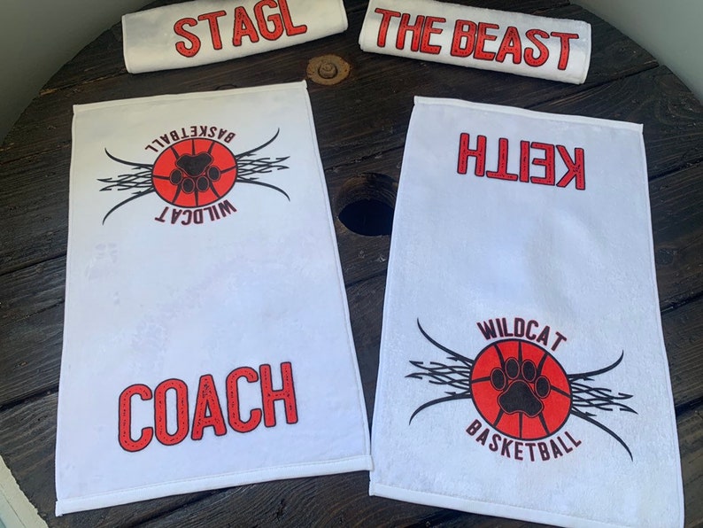 May include: Four white towels with red and black printed designs. One towel has the text "STAGL" printed in red. Another towel has the text "THE BEAST" printed in red. The third towel has the text "WILDCAT BASKETBALL" and a paw print inside a basketball. The fourth towel has the text "COACH" printed in red and the text "WILDCAT BASKETBALL" and a paw print inside a basketball.