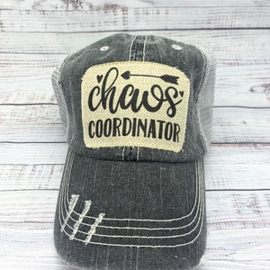 May include: A distressed black trucker hat with a mesh back and a curved brim. The hat features a patch with the words "Chaos Coordinator" in black script and an arrow design. White stitching and distressed details add to the casual style.