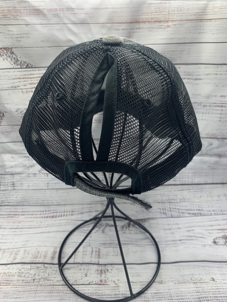 May include: Black mesh baseball cap with a ponytail hole in the back. The cap is on a black metal stand.