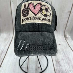 May include: A black denim trucker hat with a distressed look. The hat has a patch on the front that says "peace love soccer" with a peace sign, heart, and soccer ball graphic.