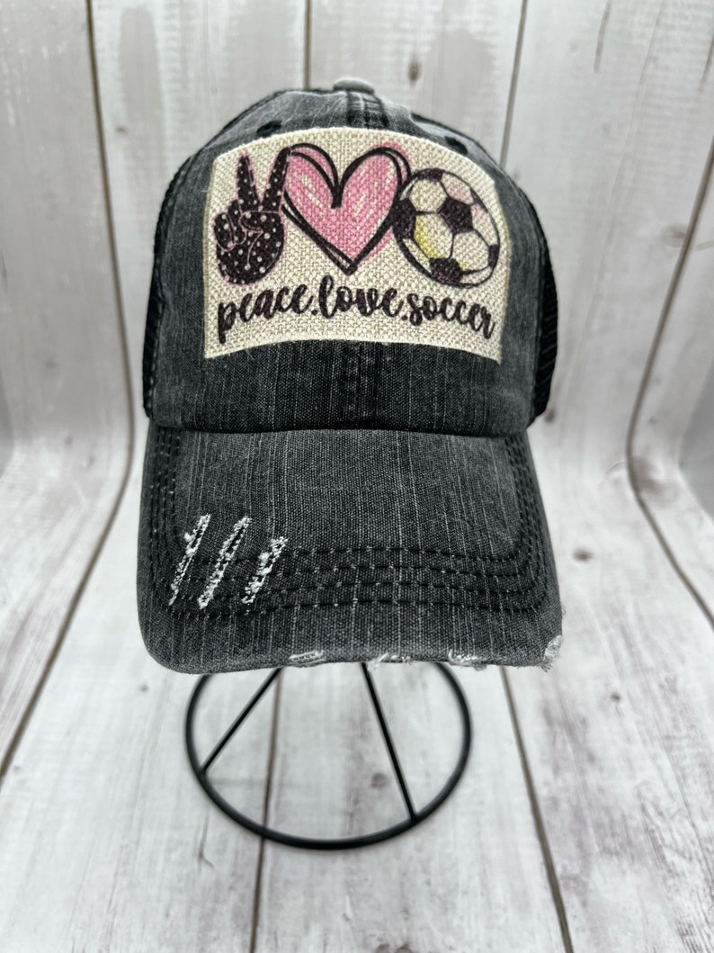May include: A black denim trucker hat with a distressed look. The hat has a patch on the front that says "peace love soccer" with a peace sign, heart, and soccer ball graphic.