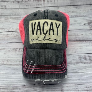 May include: A black and pink trucker hat with a distressed denim front and a pink mesh back. The hat has a white patch with the text "VACAY vibes" in black.