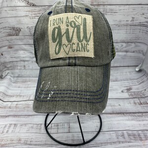 May include: A gray and blue distressed baseball cap with a patch that reads "I run a girl gang" in green lettering with a heart symbol.