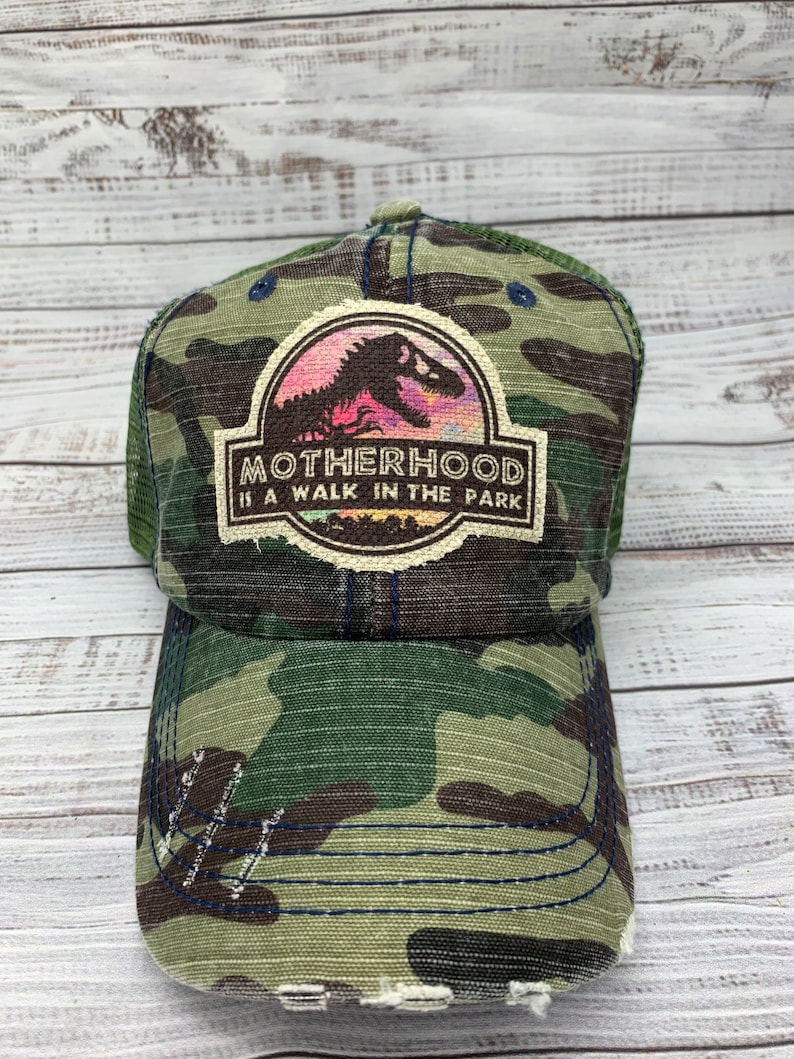 May include: A green camouflage trucker hat with a distressed look. The hat features a patch with a dinosaur silhouette and the text "MOTHERHOOD IS A WALK IN THE PARK".