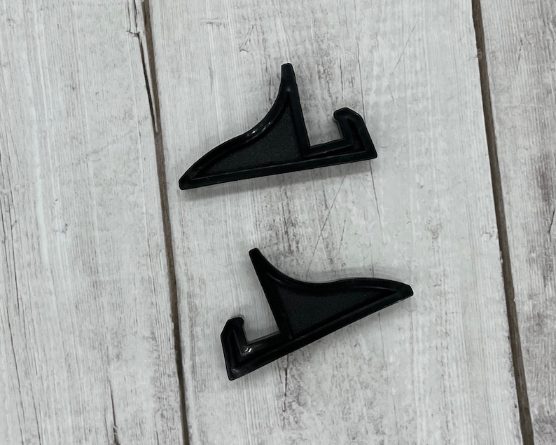 May include: Two black plastic picture frame stands. The stands are shaped like an upside down L.