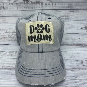 May include: A gray and blue trucker hat with a distressed look. The hat has a patch on the front that says "Dog Mom" with a paw print.
