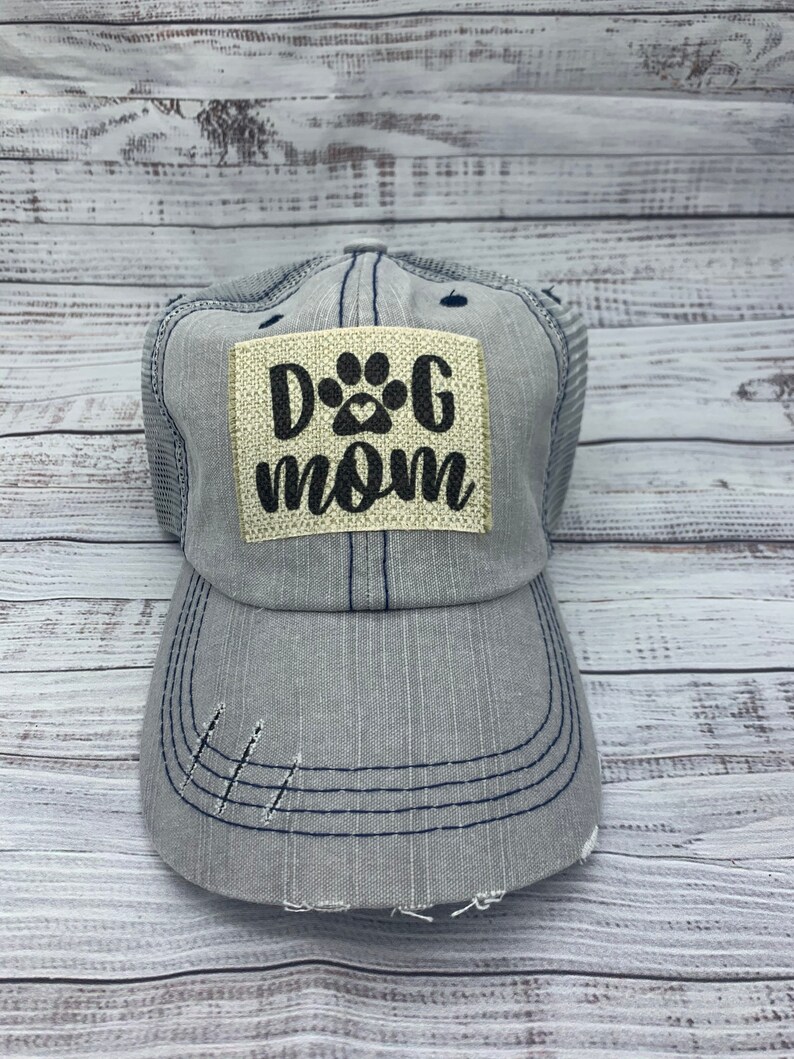 May include: A gray and blue trucker hat with a distressed look. The hat has a patch on the front that says "Dog Mom" with a paw print.