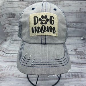 May include: A gray and blue distressed trucker hat with a beige patch that says "Dog Mom" with a paw print and heart.