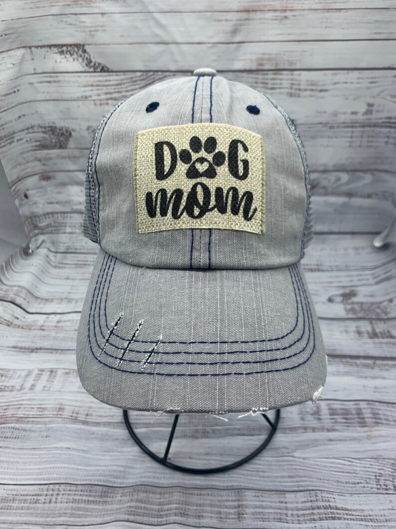 May include: A gray and blue distressed trucker hat with a beige patch that says "Dog Mom" with a paw print and heart.