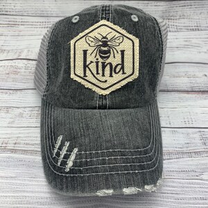 May include: Distressed black baseball cap with a mesh back. The front features a hexagon patch with a bee illustration and the word "Kind". The cap has a pre-curved bill with distressed detailing.