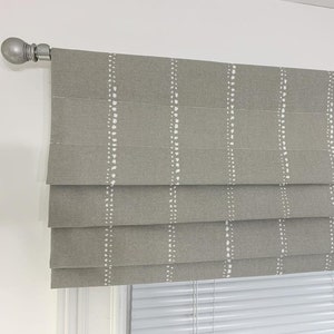 May include: Gray roman shade with a white dotted pattern. The shade is raised, revealing a white window blind underneath.