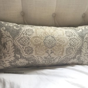 May include: A gray and beige patterned throw pillow with a floral design. The pillow is on a white bed.