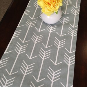 May include: A gray table runner with a white arrow pattern. A yellow flower is in a white vase on top of the runner.
