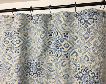 May include: A shower curtain featuring a repeating geometric pattern in shades of blue and off-white. The fabric is suspended from a black metal rod by dark metal rings, creating vertical folds across the surface of the patterned textile.