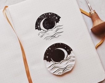 Puede incluir: Dos sellos de goma en blanco y negro con un diseño de luna creciente y olas. Los sellos están en un cuaderno blanco con un patrón de puntos.