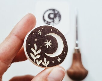 Puede incluir: Un sello de madera redondo con una luna creciente, estrellas y hojas talladas en la superficie. El sello es de color marrón oscuro con detalles blancos.