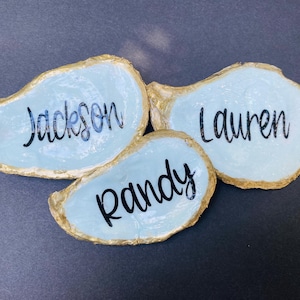 May include: Three oyster shell place cards with names Jackson, Lauren, and Randy written in black script. The shells have a light blue interior and gold-painted edges, set against a dark gray background.