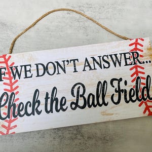 May include: A white wooden sign with red and black stitching around the edges. The sign reads "If we don't answer... Check the ball field!"
