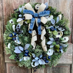 May include: A green wreath decorated with oyster shells, blue berries, and sprigs of eucalyptus. A blue and brown bow is tied at the top.